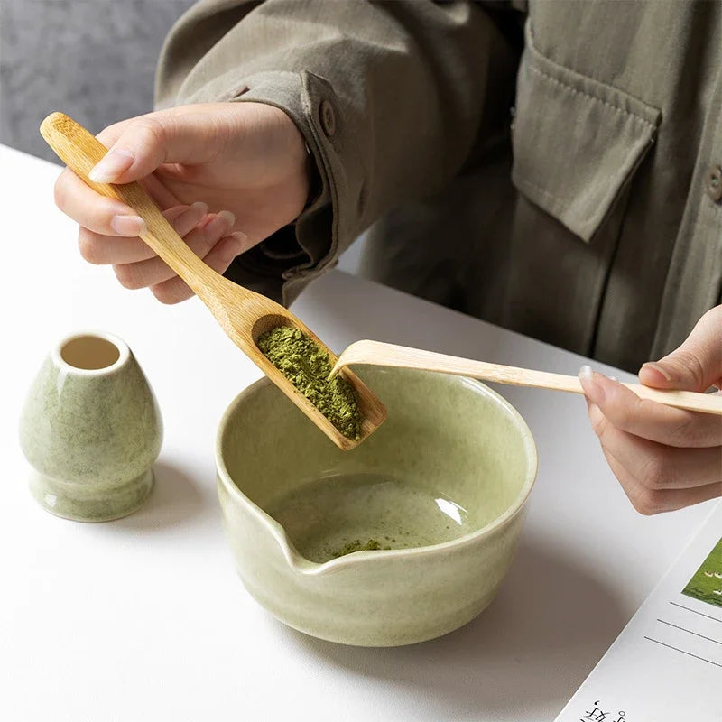 Hands spooning matcha powder into the sage bowl with the bamboo scoop
