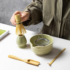 Hands holding the bamboo whisk over the sage bowl, with the sieve and scoop on the table