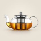 Wide glass teapot with a stainless steel lid and mesh infuser, filled with amber tea, on a warm beige background