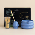 Blue matcha set on a warm beige background: speckled wavy bowl with pouring lip, whisk holder, bamboo whisk in its case and bamboo scoop, in front of its gift box