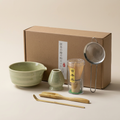 Sage matcha gift set on a warm beige background: crackle-glazed bowl with pouring lip, whisk holder, bamboo whisk, scoop and spoon, steel sieve and kraft box