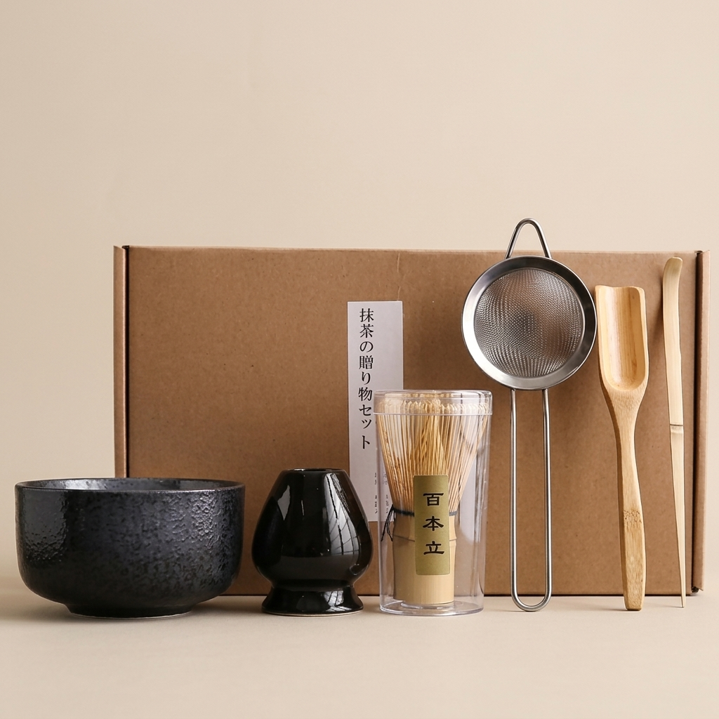 Black stone matcha set on a warm beige background: textured black bowl, glossy black whisk holder, bamboo whisk in its case, steel sieve, bamboo scoop and spoon, with the kraft box