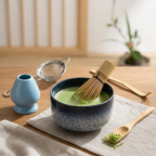 Blue speckle bowl of frothy matcha on a linen cloth by a paper screen window, with the bamboo whisk, scoop and sieve