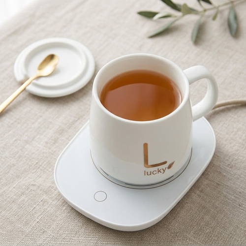 White ceramic mug of tea on its white warming plate, with a gold-tone spoon, on a light table